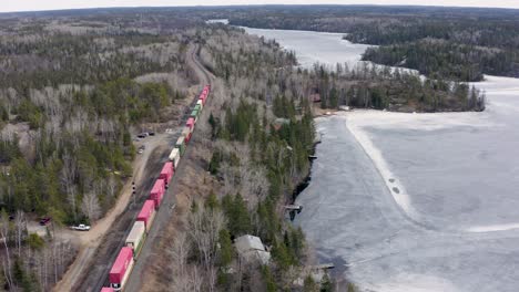 Colorful-freight-train-winds-through-the-forest-as-it-travels-along-side-a-frozen-lake