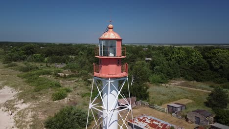 Beautiful-aerial-view-of-white-painted-steel-lighthouse-with-red-top-located-in-Pape,-Latvia-at-Baltic-sea-coastline-in-sunny-summer-day,-wide-angle-drone-shot-orbit-left