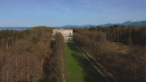 Scenic-4K-aerial-view-upon-Bavaria's-famous-castle-island-Herrenchiemsee-and-its-royal-park-at-lake-Chiemsee-in-the-rural-countryside-with-a-beautiful-blue-sky-and-the-alps-mountains-in-the-background