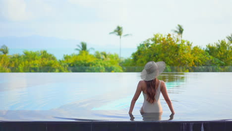 Woman-model-in-swimming-suit-and-hat-sitting-at-the-edge-of-infinity-pool-leaning-on-her-arms-on-tropical-mountains-and-green-trees-background,-daytime-slow-motion-back-view