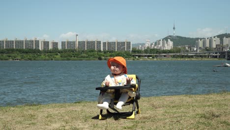 Adorable-One-Year-Old-Toddler-In-Outdoor-Baby-Seat-Under-The-Sunlight-By-The-Riverbank-In-Seoul,-South-Korea