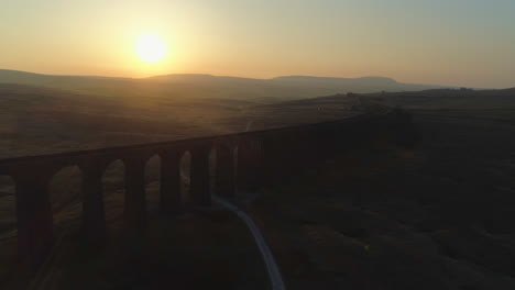 Aerial-Drone-Shot-Into-Sun-Pulling-back-From-Ribblehead-Viaduct-Train-Bridge-at-Stunning-Sunrise-in-Summer-in-Yorkshire-Dales-England-UK-with-Hills-in-Background