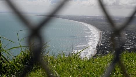 Grassy-edge-of-a-mountain-towering-above-a-oceanfront-town-with-chain-link-fence-in-foreground