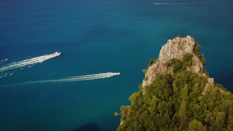 Perfectly-composed-drone-orbit-shot-of-coastal-rock-formation-with-trees,-small-cruise-ship-and-speedboat-sailing-vivid-blue-waters-in-the-background