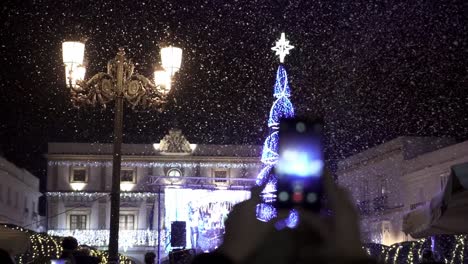 Slow-motion-handheld-shot-of-a-christmas-party-in-spain-in-medina-sidonia-in-cadiz-spain-with-a-lighted-christmas-tree-with-a-star-on-top-while-a-person-with-a-smartphone-is-filming-with-snowfall