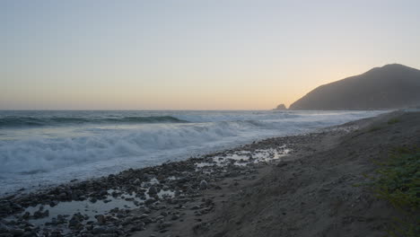 Stationary-shot-of-waves-crashing-against-the-rocky-shore-of-Mondo's-Beach-with-sunsetting-behind-a-mountain-located-in-Southern-California