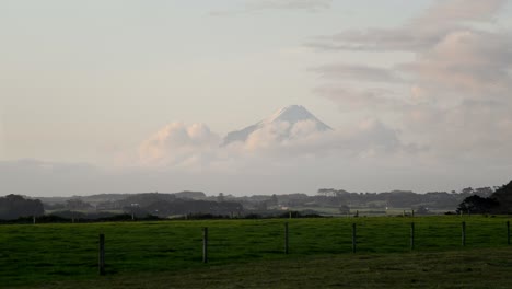 Majestic-Mount-Taranaki-peeking-out-the-soft-evening-clouds-illuminated-by-golden-hour-light-with-farm-land-in-foreground