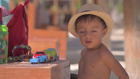 Junges-Lateinisches-Kleinkind-Mit-Hut,-Das-Mit-Seinem-Auto-Spielt,-Spielt-Auf-Einer-Bank-Mit-Sand-Am-Strand