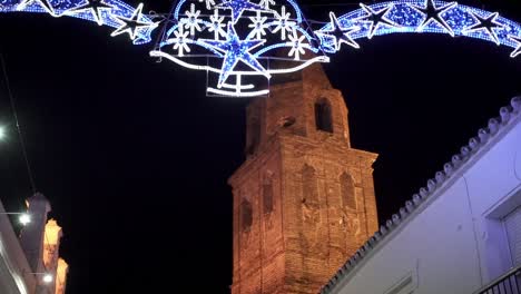 Slow-motion-tilt-up-shot-of-an-entrance-arch-decorated-for-christmas-with-lights,-glowing-snowflakes-and-star-overlooking-a-church-tower-in-medina-sidonia-in-spain-cadiz