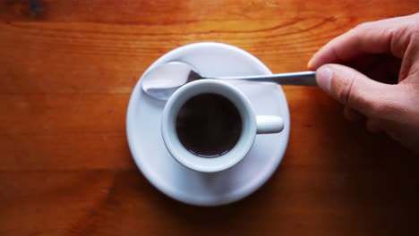 Hand-is-putting-spoon-next-to-a-hot-espresso-cup,-standing-on-a-brown-wooden-table