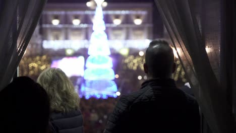 Slow-motion-handheld-panning-shot-of-people-looking-out-of-an-open-window-with-curtains-at-a-christmas-festival-in-medina-sidonia-in-cadiz-spain-in-the-square-a-glowing-christmas-tree-in-blue