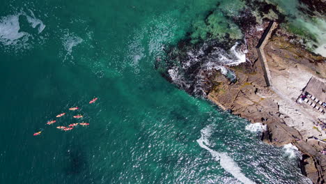 Top-down-view-of-bright-kayaks-paddling-away-from-rocky-coastline