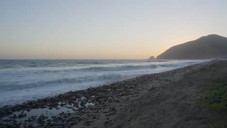 stationary-shot-of-waves-rolling-into-the-shores-of-Mondo's-Beach-with-sun-setting-behind-mountain-in-the-background-located-in-Southern-California
