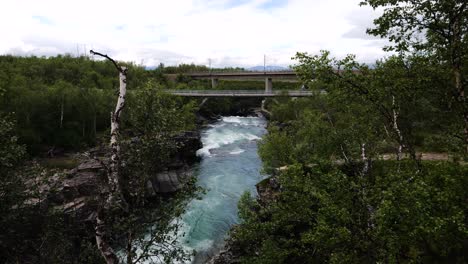 Mit-Blick-Auf-Fließendes-Flusswasser-In-Schweden,-Gesehen-Hinter-Dünnen-Kahlen-Bäumen-Mit-Zwei-Brücken-Im-Hintergrund