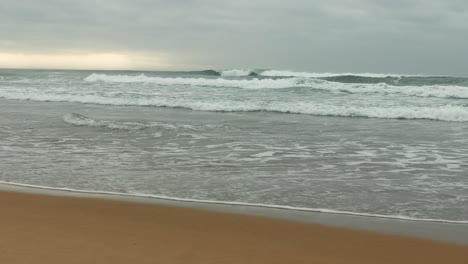 Waves-breaking-onto-sandy-beach,-cloudy-skies-in-background