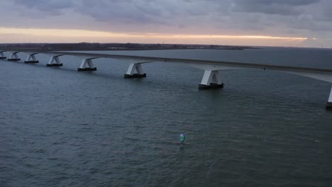 Luftaufnahme-Einer-Längsten-Brücke-Von-Zeelandbrug-In-Holland-Mit-Einem-Surfer-Auf-Einem-Tragflächenprofil-Mit-Blauem-Flügelwind,-Der-Darunter-Foliert