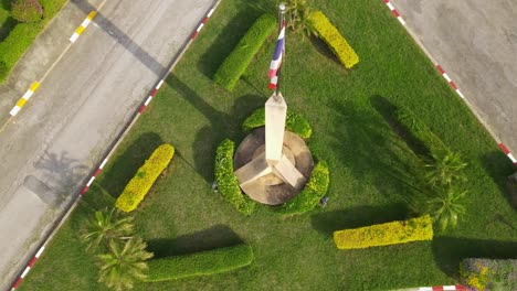 Bandera-Nacional-Tailandesa-En-Un-Poste-Con-Pedestal,-Imágenes-Aéreas-Descendentes-De-La-Bandera-Tailandesa-Que-Revelan-El-Pedestal-Y-La-Hierba