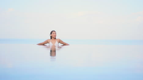 Attractive-Asian-woman-with-flower-behind-ear-relaxing-in-resort-infinity-pool-with-reflection-on-surface-of-water