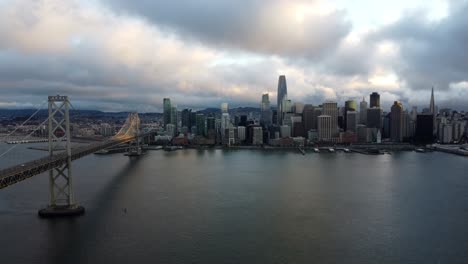 4K-30fps-Aerial-Drone-Footage-of-San-Francisco-City-Skyline-and-the-Oakland-Bay-Bridge---Cars-Travel-Along-the-Highway,-Stormy-Cumulus-Clouds,-Calm-Bay-Water,-Sunset-Reflecting-off-Waves,-Landscape