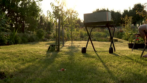 Barbecue-grill-with-fire-on-nature,-grill-in-garden-and-a-man-sitting-on-a-chair,-a-dog-is-playing-in-sun-light-and-trees-in-garden
