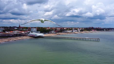 Seagull-flying-towards-the-drone-as-it-was-capturing-a-revers-footage-of-a-pier,-beachfront,-townhouses,-and-a-gorgeous-British-sky-with-huge-rain-clouds