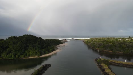High-static-drone-view-of-a-large-rainbow-on-a-sunny-day-with-scenic-outlook-towards-a-river-mouth-leading-out-to-a-rough-ocean
