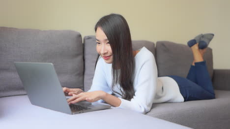 Close-up-of-an-attractive-young-woman-in-jeans-and-a-sweatshirt-working-on-her-laptop-while-lying-on-her-stomach-on-the-sofa
