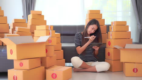 A-young-woman-sits-among-shipping-boxes-doing-inventory-and-recording-them-on-a-clipboard