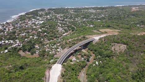 Aerial-view-of-a-concrete-bridge,-with-cars-passing-by-and-a-beautiful-view-of-the-beach-in-the-background-with-waves-crashing-all-the-way-to-the-beach