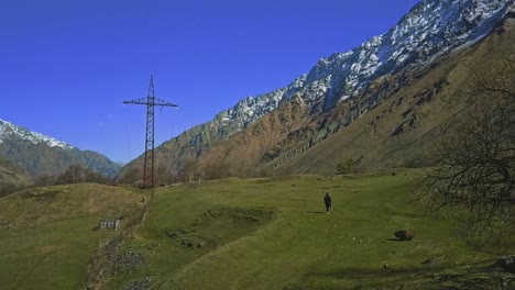 Einsamer-Wanderer-In-Den-Bergen-Mit-Poncho-Und-Stock,-Hochspannungsleitung,-Bäume,-Schneebedeckter-Berg,-Luftbild