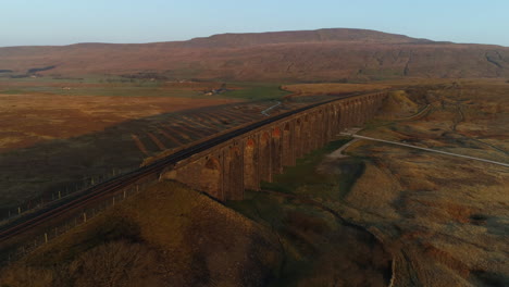 Slow-Aerial-Drone-Shot-of-Ribblehead-Viaduct-Train-Bridge-with-Long-Shadows-at-Stunning-Sunrise-in-Summer-in-Yorkshire-Dales-England-UK-with-3-Peaks-Whernside-Mountain-in-Background