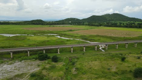 Angled-aerial-sliding-footage-towards-the-left-revealing-the-elevated-railways,-farmland-with-cattle,-mountains-and-lake-in-the-horizon,-Muak-Klek,-Saraburi,-Thailand