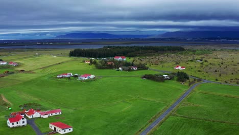 Panoramablick-Auf-Die-Grüne-Wiese-Auf-Dem-Friedhof-Von-Gardakirkjugardur-Mit-Unbefestigter-Straße,-Einigen-Häusern-Und-Einer-Kirche-In-Der-Ferne-In-Der-Nähe-Von-Reykjavik,-Island