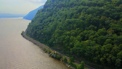 Aerial-drone-footage-of-freight-train-tracks-along-the-Hudson-River-in-New-York's-Hudson-Valley-during-early-autumn-with-mountains-and-a-river-and-forests
