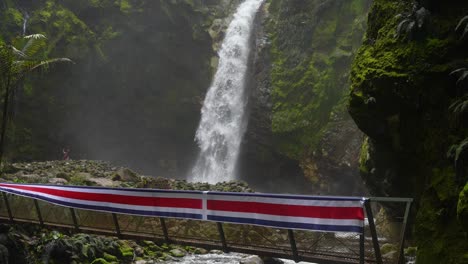 Wasserfall-Neben-Brücke-Mit-Costa-Rica-Flagge-Oben-Auf-Berg-Mit-Fluss