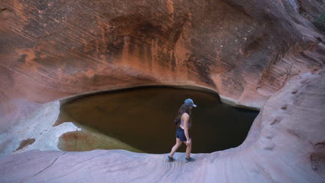 Young-Female-Hiker-Walking-on-Sandstone-Cliff-Above-Rainwater-Pond-in-Wilderness-of-Utah,-St