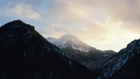 Descending-aerial-drone-shot-of-a-winter-landscape-of-Mount-Timpanogos-in-the-background-surrounded-by-a-pine-tree-forest-during-sunset-from-Tibble-Fork-Reservoir-in-American-Fork-Canyon,-Utah