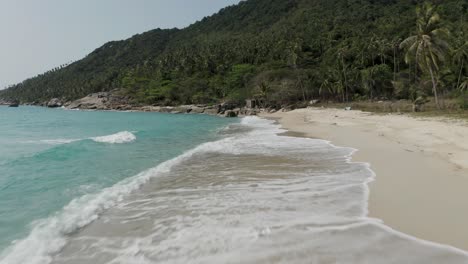Waves-breaking-on-beautiful-white-sand-beach-in-Koh-Phangan,-low-aerial