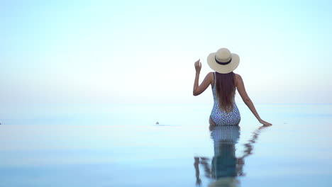 Back-view-of-Woman-in-swimsuit-and-Hat-sitting-on-the-edge-of-rooftop-infinity-pool-with-body-reflection-in-water-and-sunset-sky,-vacation-template