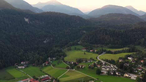 Aerial-view-over-a-summer-forest-during-sunrise-with-mountain-landscape-in-the-Austrian-Alps,-small-town-with-wooden-houses-in-the-background