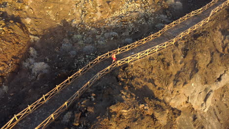 Woman-in-a-red-dress-walks-along-a-path-with-a-railing-on-a-lava-island-at-golden-sunset,-Charco-de-los-Sargos---drone-shot