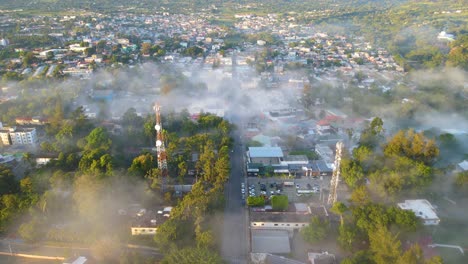 Jarabacoa-aerial-view,-city-covered-with-fog-in-the-morning,-stunning-environment-of-a-small-town-in-the-Dominican-Republic-with-trees-and-mountains-behind-the-city