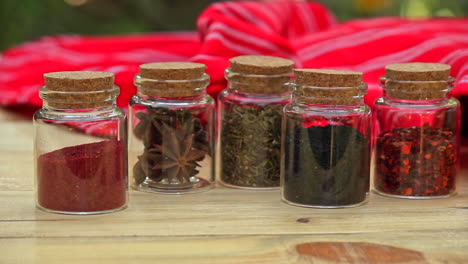 Selection-of-Jars-with-dried-spices-laid-out-in-a-row-on-wooden-table-with-red-cloth-in-the-background