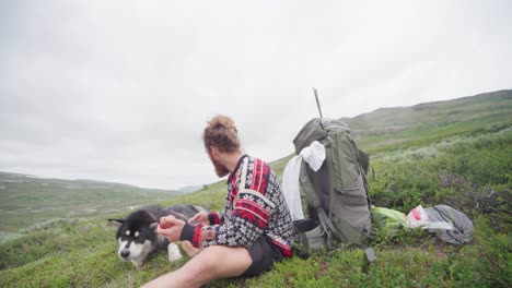 Handsome-Man-With-Beard-Eating-Fruit-With-His-Alaskan-Malamute-On-A-Grassy-Mountain-Peak