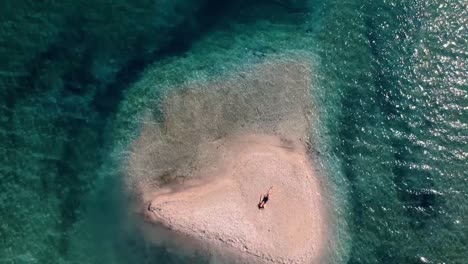 Top-down-Shot-Of-A-Beautiful-Caucasian-Woman-Sunbathing-On-A-Small-Island-In-Italy-Surrounded-By-Bright-Blue-Sea-On-A-Summer-Day---aerial