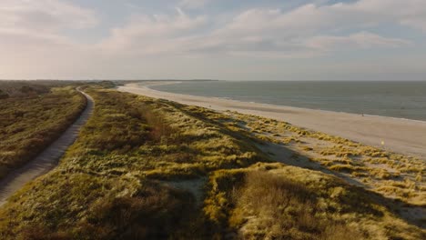 Long-aerial-shot-flying-along-a-path-through-grassy-dunes-along-an-empty-beach-at-sunset