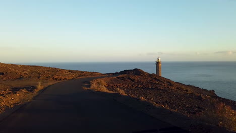 Driving-along-the-road-at-sunset-to-the-lighthouse-towering-on-the-rocky-coast-of-Heirro-Island,-Faro-de-la-Orchilla