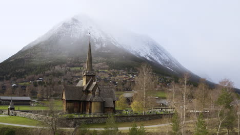 Vista-Escénica-De-La-Iglesia-De-Lom-Stave-Contra-Una-Enorme-Montaña-Nevada-En-Invierno-En-Noruega