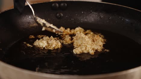 Chef-using-a-spoon-to-spread-out-the-chopped-garlic,-making-sure-it-is-evenly-covered-in-the-cooking-oil,-slowly-frying-garlic-in-the-shallow-frying-pan-in-low-heat,-close-up-shot