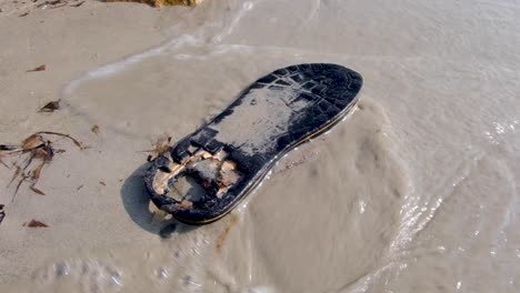 A-single-lost-and-partially-decomposed-black-flip-flop-on-a-sandy-beach-with-waves-washing-over-it-polluting-and-littering-on-a-remote-tropical-island